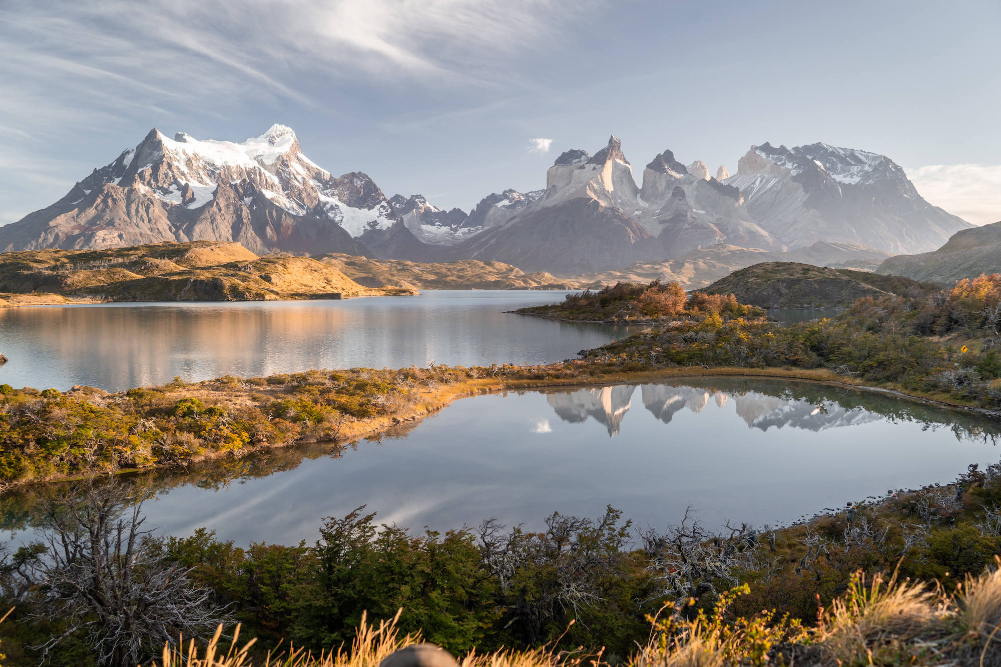 Calm waters act as a perfect mirror, doubling the magnitude of the snow-capped peaks in the distance. The golden hue of the shoreline vegetation emphasises the stillness of a windless autumn morning.