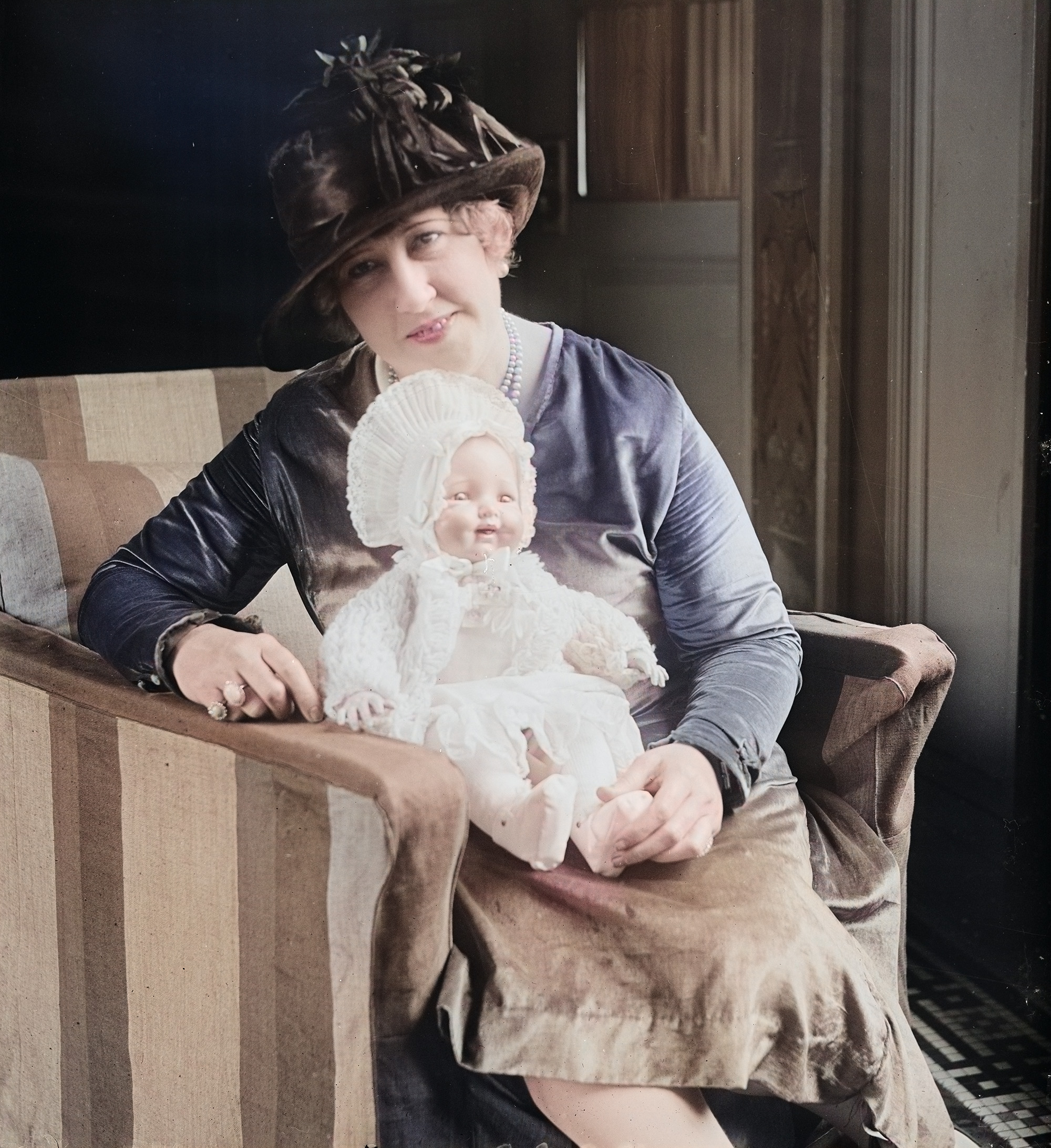 A colorized historical photograph shows prolific inventor Beulah Louise Henry seated in a striped armchair. She is wearing a brown velvet hat with feathered accents, a purple satin dress, and a pearl necklace. Resting on her lap is her invention, a "washable air baby" doll, which is dressed in an intricate white bonnet and lace outfit.