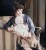 A colorized historical photograph shows prolific inventor Beulah Louise Henry seated in a striped armchair. She is wearing a brown velvet hat with feathered accents, a purple satin dress, and a pearl necklace. Resting on her lap is her invention, a