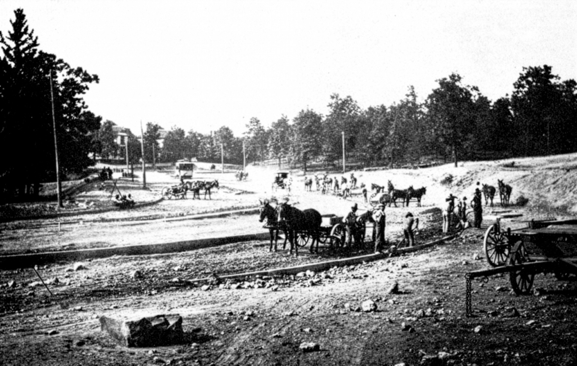 A historical black-and-white photograph showing the early construction of Highland Avenue. The scene features several horse-drawn wagons and teams of horses working on a wide, dirt roadbed. Laborers are scattered across the landscape, which is lined with sparse trees and a few distant houses under a bright sky.