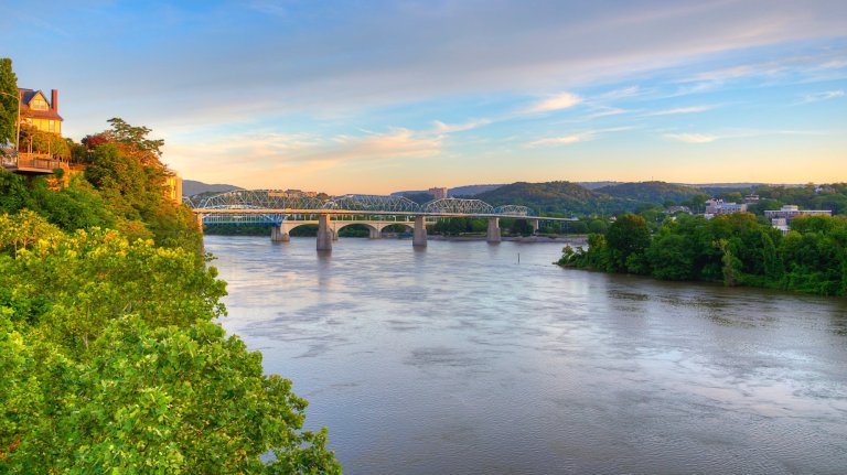 A serene Tennessee River flows beneath the Walnut Street Bridge during a picturesque sunrise, with lush green trees lining its banks. Houses are perched on a hill to the left, overlooking the tranquil waters. Walnut Street Bridge Chattanooga, TN Photo taken on May 12, 2024