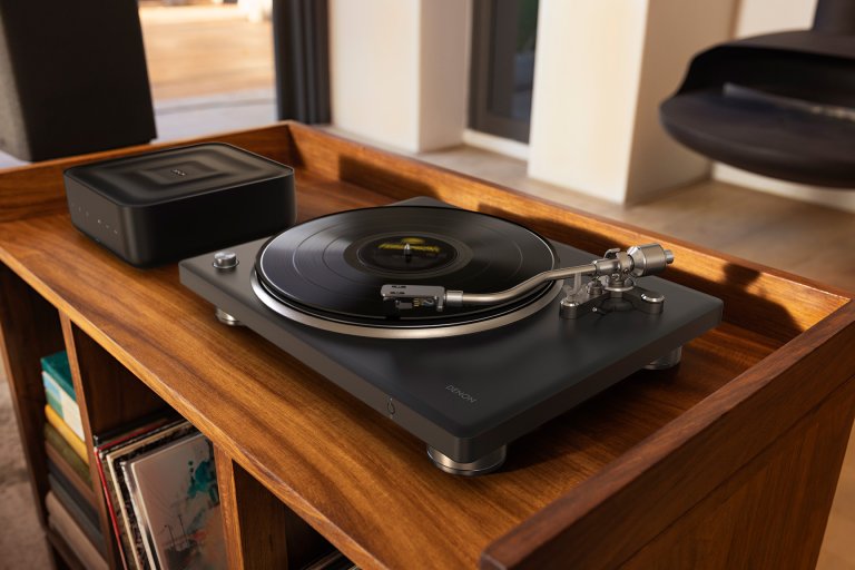 A black two-tone Denon DP-500BT turntable sitting next to a Denon Home Amp on a brown wood record stand