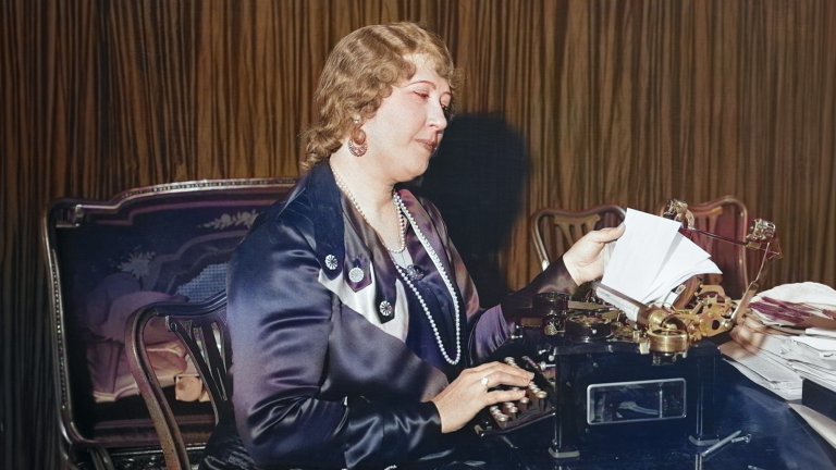 A colorized historical photograph of prolific inventor Beulah Louise Henry, known as "Lady Edison," seated at a desk. She is dressed in a formal purple satin dress and pearls, operating a complex, specialized typewriter of her own design. She is looking down at a sheet of paper she is feeding into the machine.