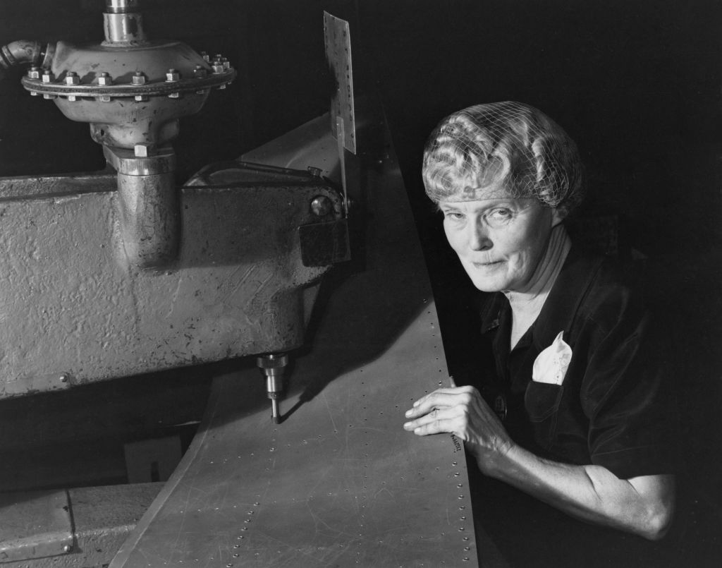 A black-and-white historical photograph of Mabel Stark, a famous tiger trainer, working in a war plant during World War II. Stark is shown wearing a hairnet and a dark work shirt, operating a large industrial machine used for precision drilling or riveting on a long metal aircraft component. She is looking directly at the camera with a focused expression while her hand rests near a row of drilled holes on the metal sheet.