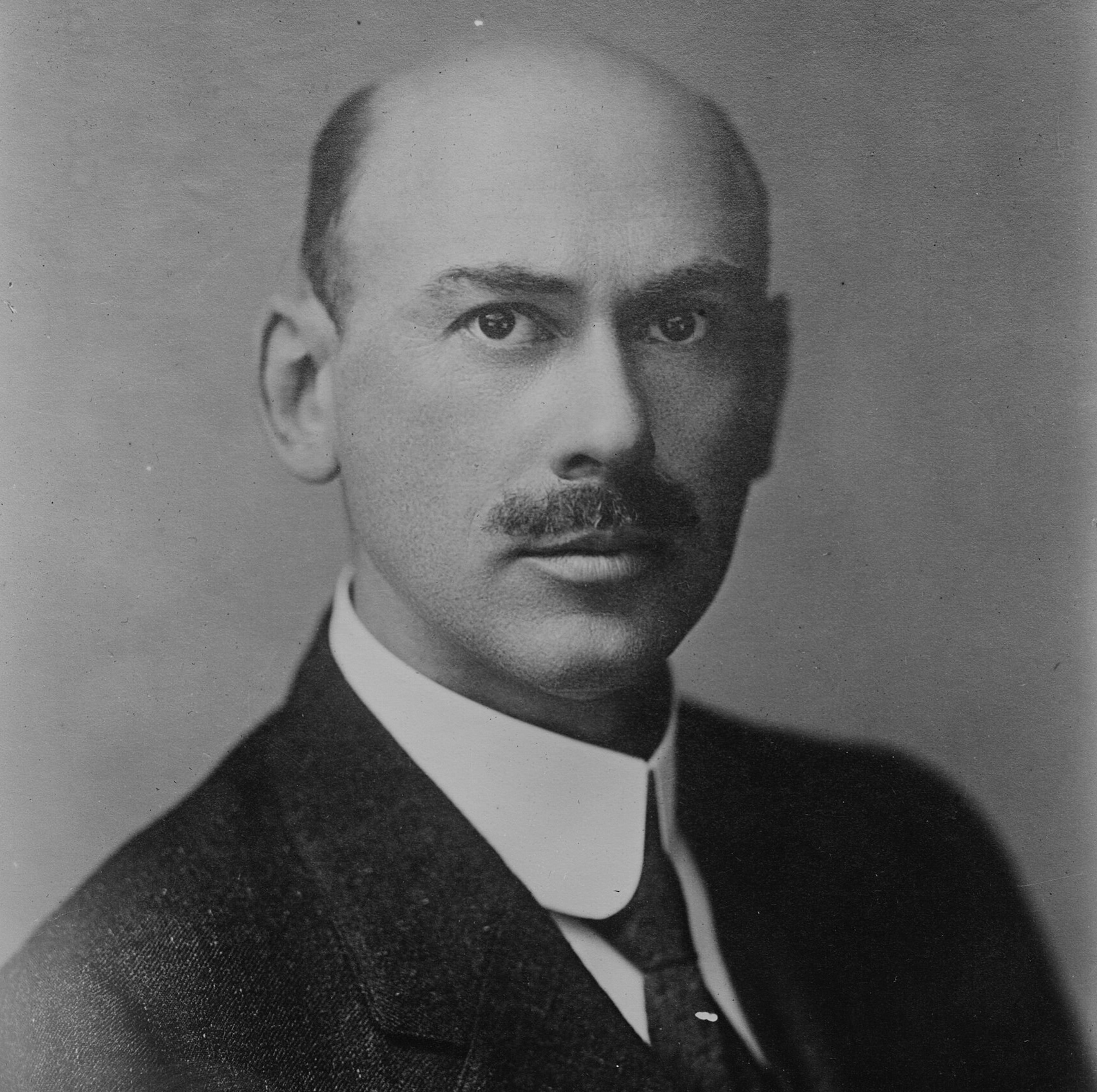 A black-and-white formal portrait of Robert Goddard, widely considered the father of modern rocketry. He is a bald man with a dark mustache, wearing a suit jacket, a high-collared white shirt, and a dark necktie.