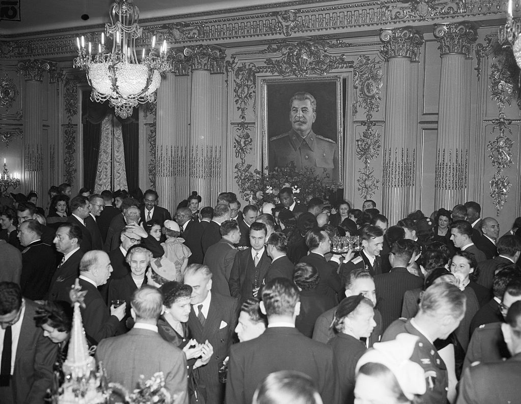 A black-and-white historical photograph of a crowded, formal party held at the Soviet Embassy in Washington, D.C.. Dozens of guests in suits and cocktail dresses fill an ornate ballroom featuring gilded columns and a large crystal chandelier. Dominating the center wall is a large framed portrait of Joseph Stalin in military uniform, overlooking the gathered crowd.