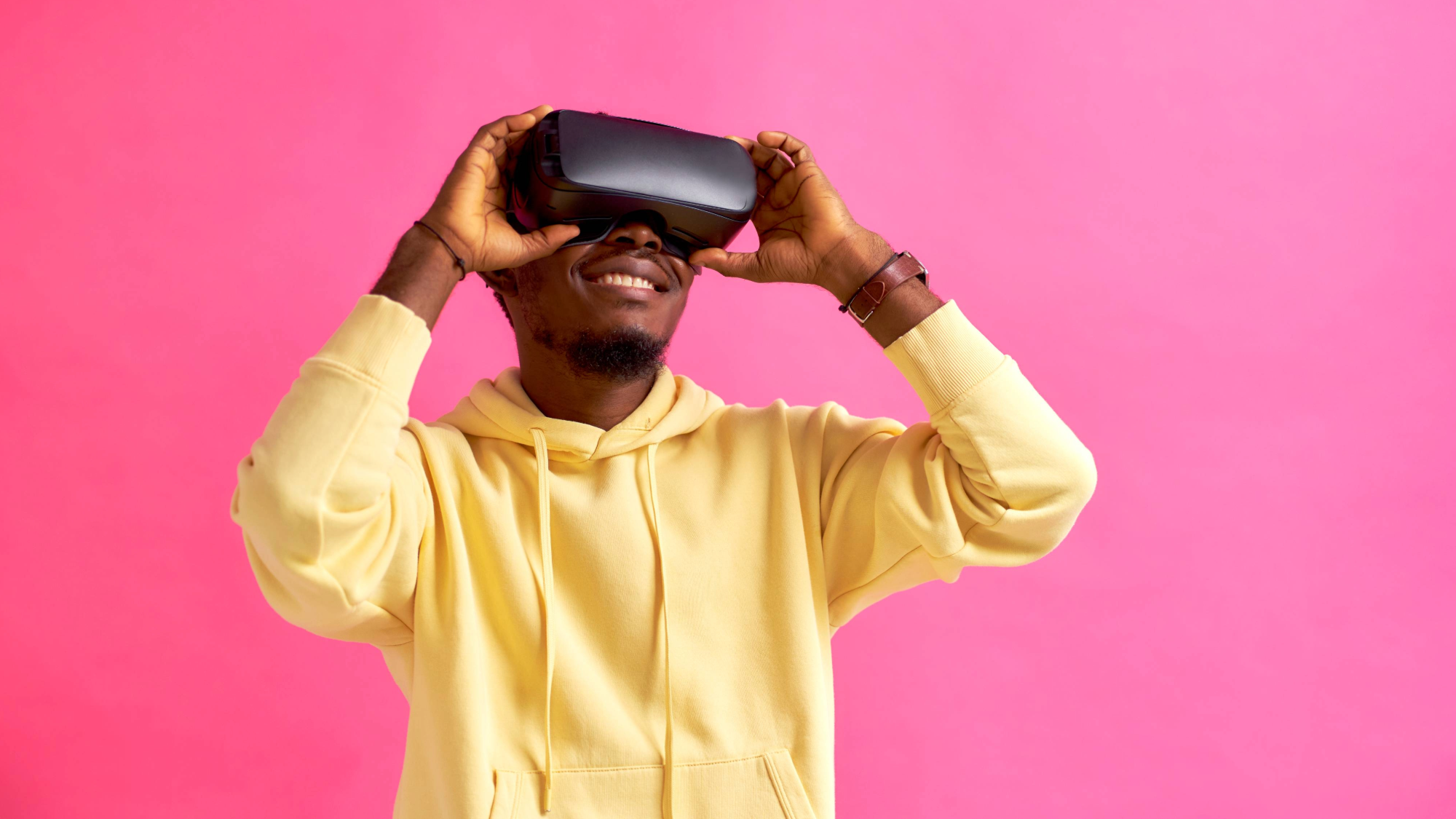 a smiling man in a yellow hooded sweatshirt wears a virtual reality headset over his eyes