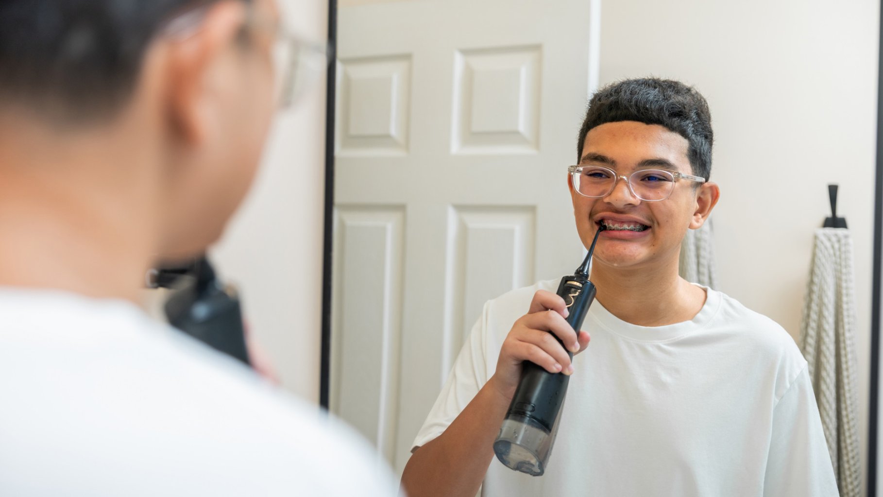 Teenage boy with dark curly hair with braces exercising good dental using a water pick while looking at his reflection in a bathroom mirror.