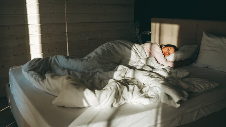 A wide-angle photograph shows a woman sleeping on her side in a large bed, tucked under a heavy, rumpled white duvet. Her head rests on a white pillow, and her face is partially illuminated by warm morning sunlight streaming through the slats of a window. The room has a minimalist aesthetic with light-colored wooden paneled walls and soft, high-contrast lighting that creates long shadows across the bed.