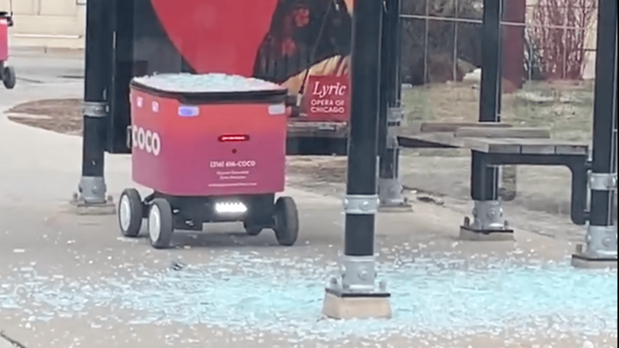 a red delivery robot that crashed into a bus shelter. broken glass is all over the side walk