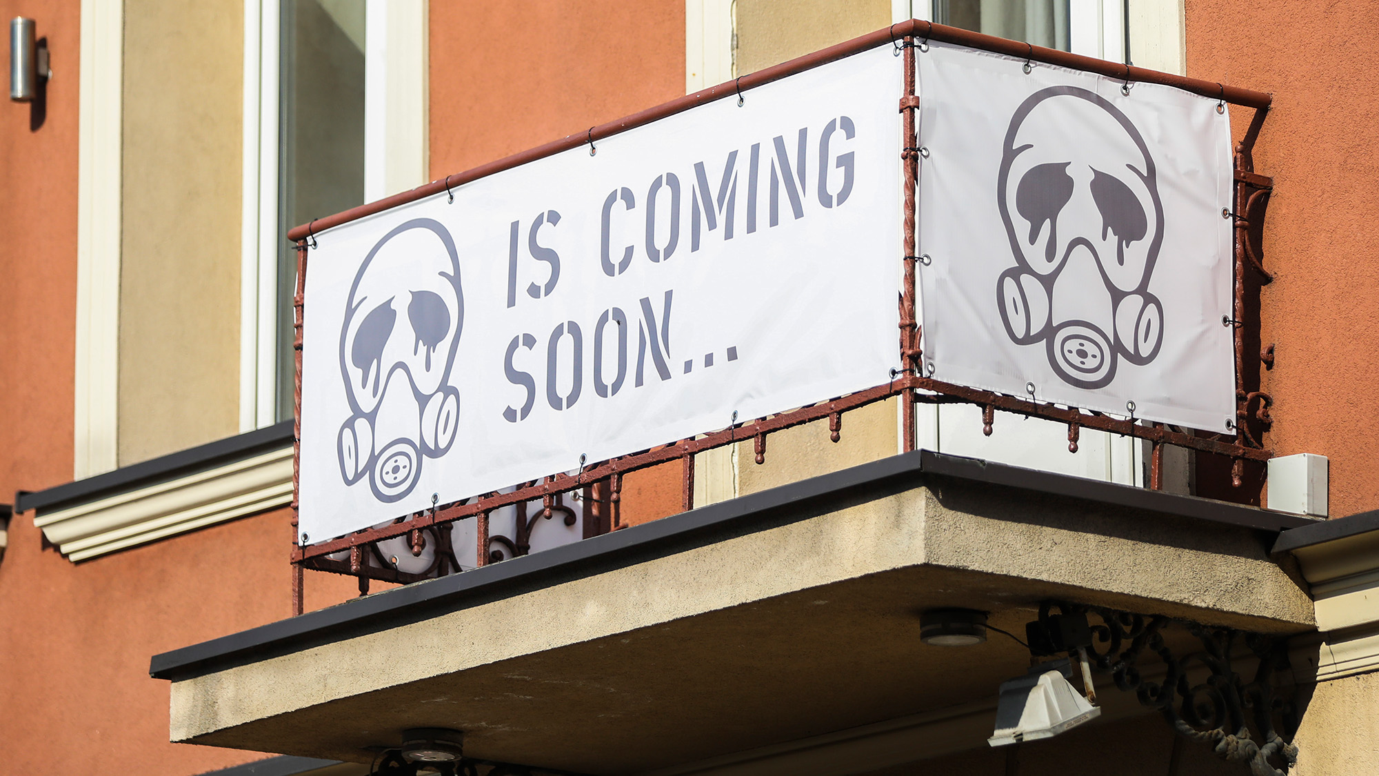 A photograph of an orange apartment building balcony featuring two large white banners attached to the railing. The front-facing banner displays the text "IS COMING SOON..." in a grey stencil-style font, flanked on the left by a grey graphic of a gas mask with liquid dripping from its eye lenses. A second, identical gas mask graphic is visible on the side banner.