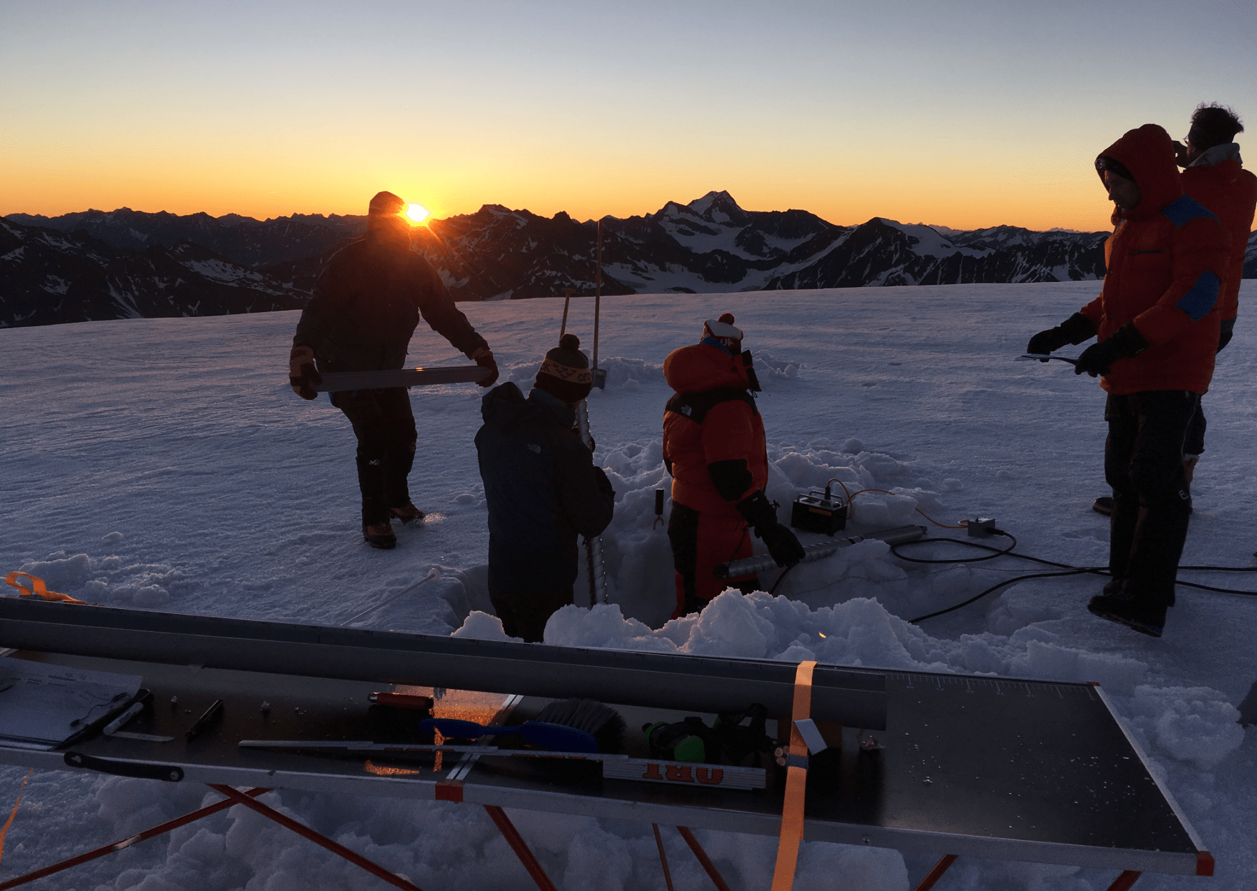 scientists on top of a glacier are drilling into the ice as the sun sets