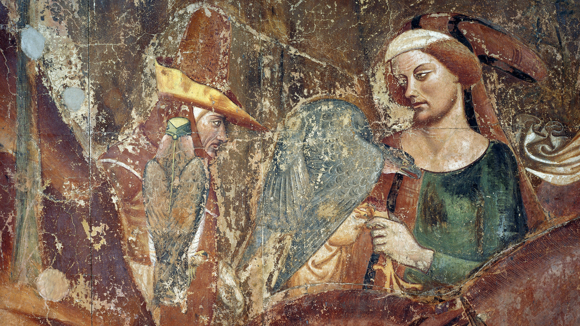 An aged and weathered medieval fresco depicts two figures engaged in falconry. On the right, a woman with auburn hair wearing a green and red bodice and a white headpiece holds a large, grey-feathered hawk or falcon. To the left, a second figure in a tall, pointed yellow and red hat observes, with a smaller brown bird of prey perched nearby. The artwork shows significant signs of age, including fading, cracks, and surface wear across the earthy-toned background.