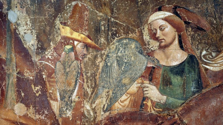 An aged and weathered medieval fresco depicts two figures engaged in falconry. On the right, a woman with auburn hair wearing a green and red bodice and a white headpiece holds a large, grey-feathered hawk or falcon. To the left, a second figure in a tall, pointed yellow and red hat observes, with a smaller brown bird of prey perched nearby. The artwork shows significant signs of age, including fading, cracks, and surface wear across the earthy-toned background.