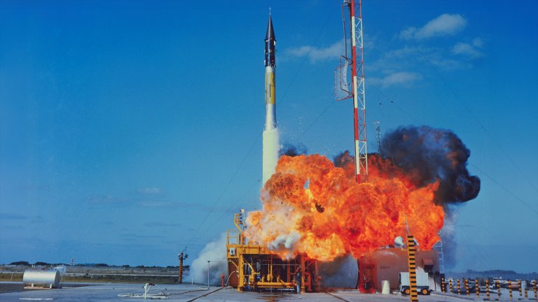 A color photograph captures the explosive failure of a Vanguard TV-3 rocket on its launchpad. A massive ball of bright orange fire and billowing black smoke erupts from the base of the slender, silver and black rocket. The rocket remains momentarily upright amidst the blast, positioned next to a tall red and white gantry tower under a clear blue sky.
