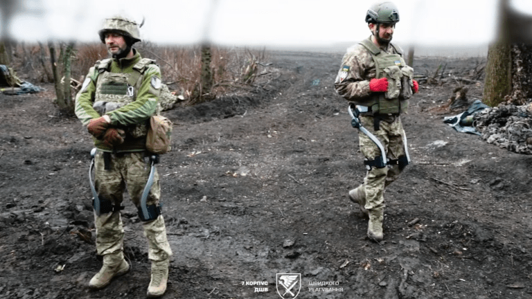 two soldiers standing in a dirty field wearing exoskeletons on their legs
