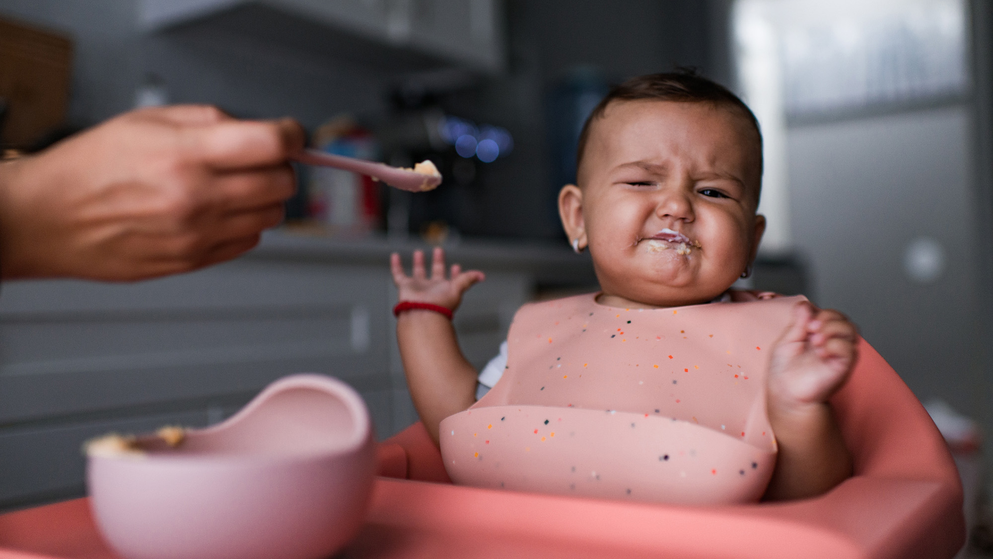 A close-up photograph of a baby sitting in a pink high chair, wearing a pink silicone bib. The baby has a distressed facial expression with eyes squinted and mouth tightly shut, appearing to refuse a spoonful of food being held out by an adult's hand. A small pink bowl sits on the tray in the foreground.