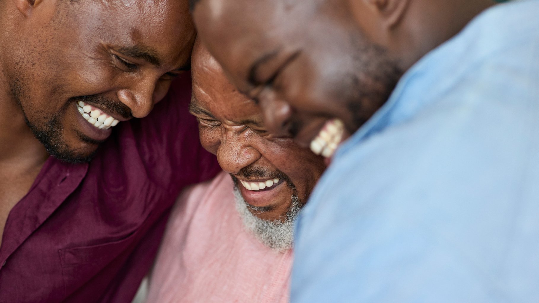A tight, candid close-up captures three Black men of different generations leaning their heads together in shared laughter. The man in the center is older with a grey beard, flanked by two younger men. All three have their eyes closed or squinted with joy, showcasing genuine, broad smiles and visible teeth. The image has a warm, intimate feel, emphasizing a strong emotional connection.