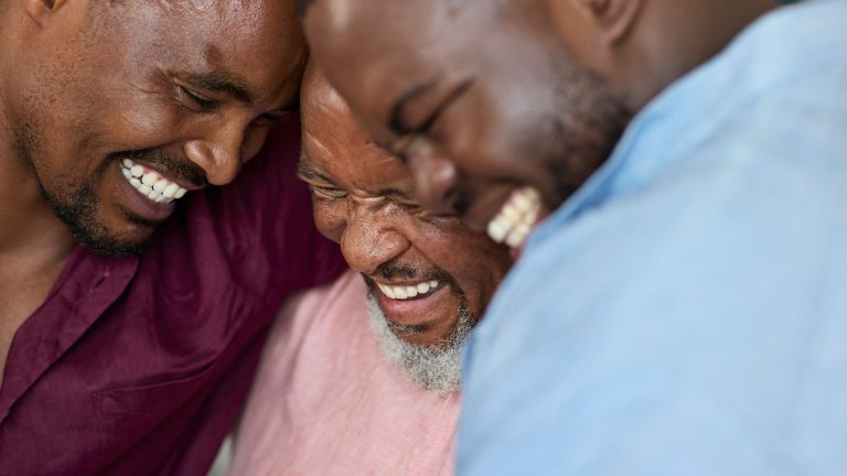 A tight, candid close-up captures three Black men of different generations leaning their heads together in shared laughter. The man in the center is older with a grey beard, flanked by two younger men. All three have their eyes closed or squinted with joy, showcasing genuine, broad smiles and visible teeth. The image has a warm, intimate feel, emphasizing a strong emotional connection.
