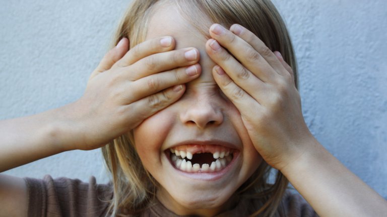 A close-up photograph of a young child with blonde hair smiling broadly, revealing several missing front baby teeth. The child is playfully covering their eyes with both hands, with a plain grey wall in the background.