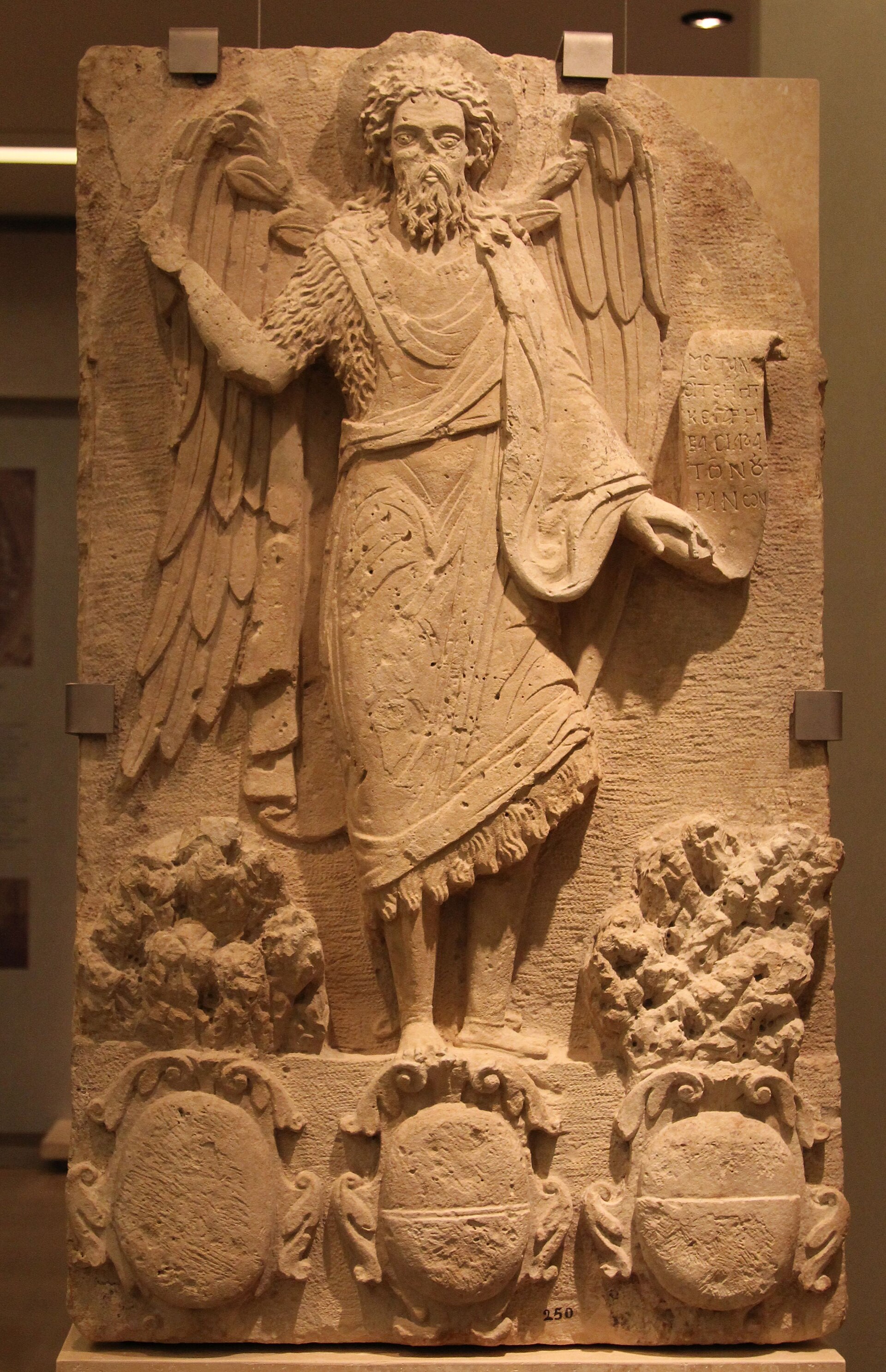 A vertical photograph shows an early 17th-century limestone panel with St. John the Baptist in relief. The carving depicts the saint as an "Angel of the Desert," featuring large wings and wearing a textured camel-hair garment. He holds an unrolled scroll with Greek inscriptions in his left hand, while his right hand is raised in a gesture of blessing. At the base of the panel are three decorative shields or coats of arms, and the figure stands amidst stylized rocky terrain.