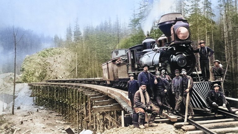 A colorized historical photograph captures a group of railroad workers posing with a large steam locomotive on a wooden trestle bridge. The workers, a diverse group including several men in rough work clothes and hats, are clustered around the front of the black engine, which features a prominent cone-shaped smokestack. The trestle bridge curves through a dense evergreen forest with a rocky hillside in the background, illustrating the challenging terrain of early American railroad construction.