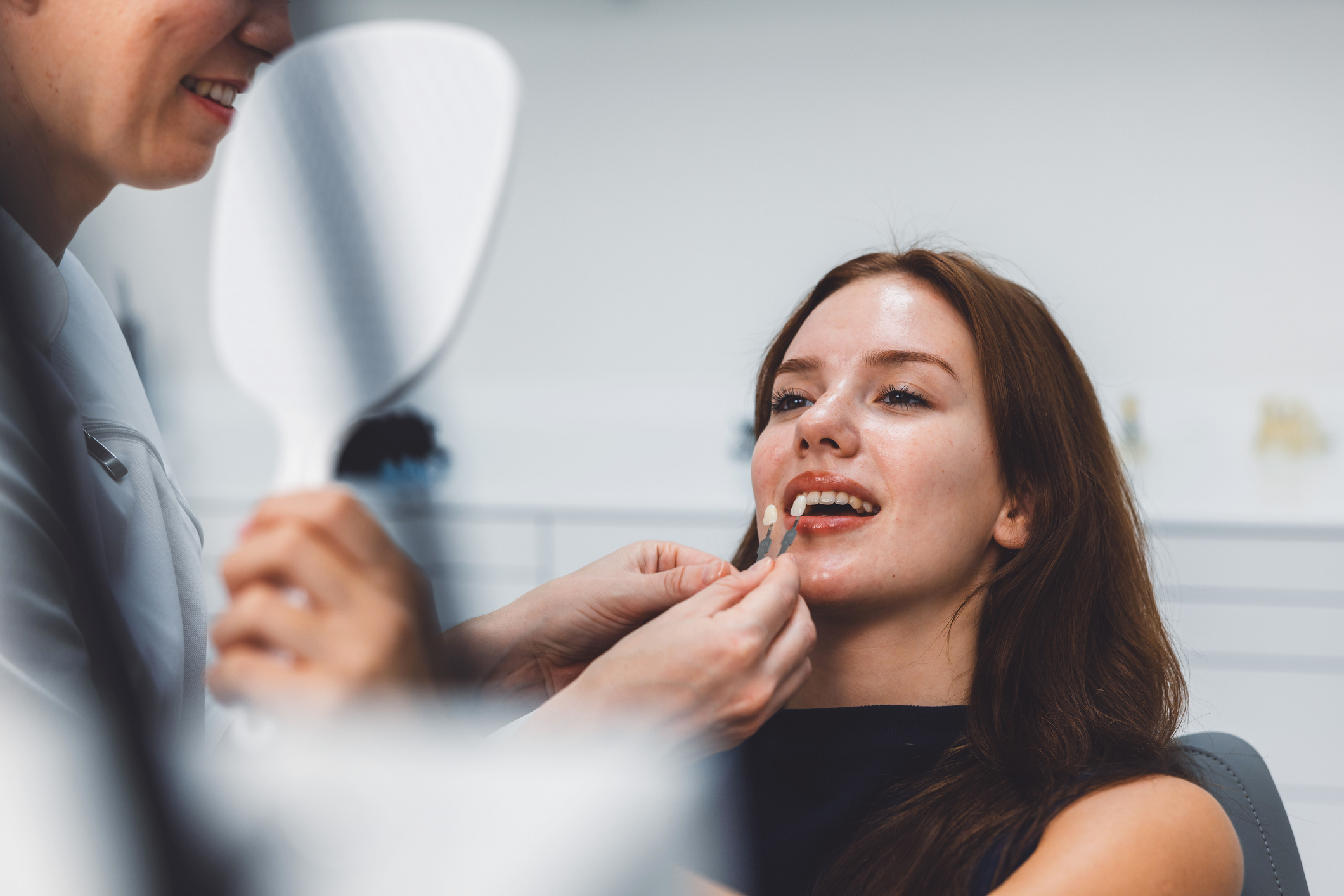 A dental professional is fitting a dental veneer on a smiling patient in a modern dental clinic. The red-headed, young female patient looks pleased as she examines her smile in a handheld mirror.