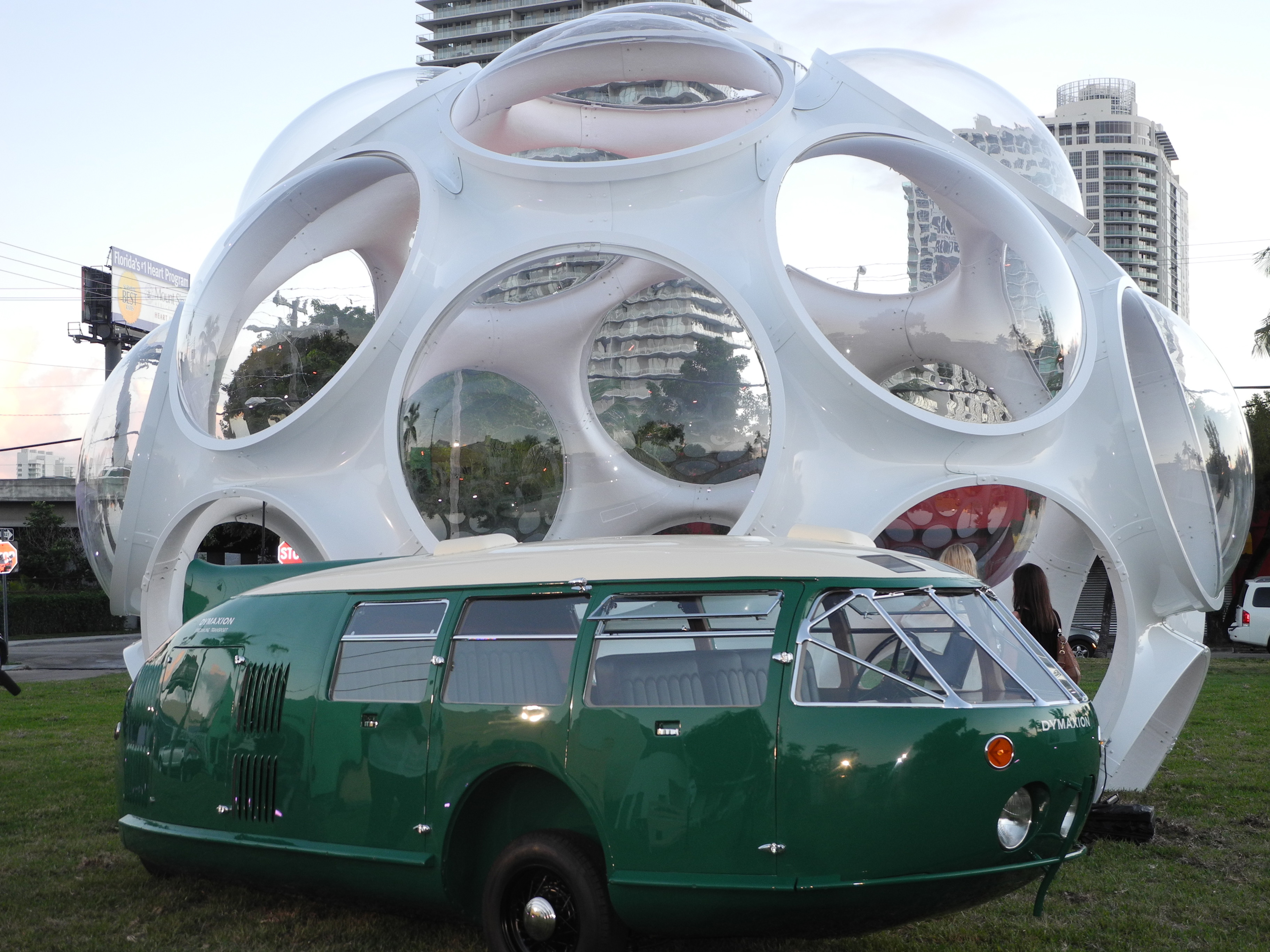 A green teardrop-shaped vintage car is parked in front of a futuristic-looking sphere.