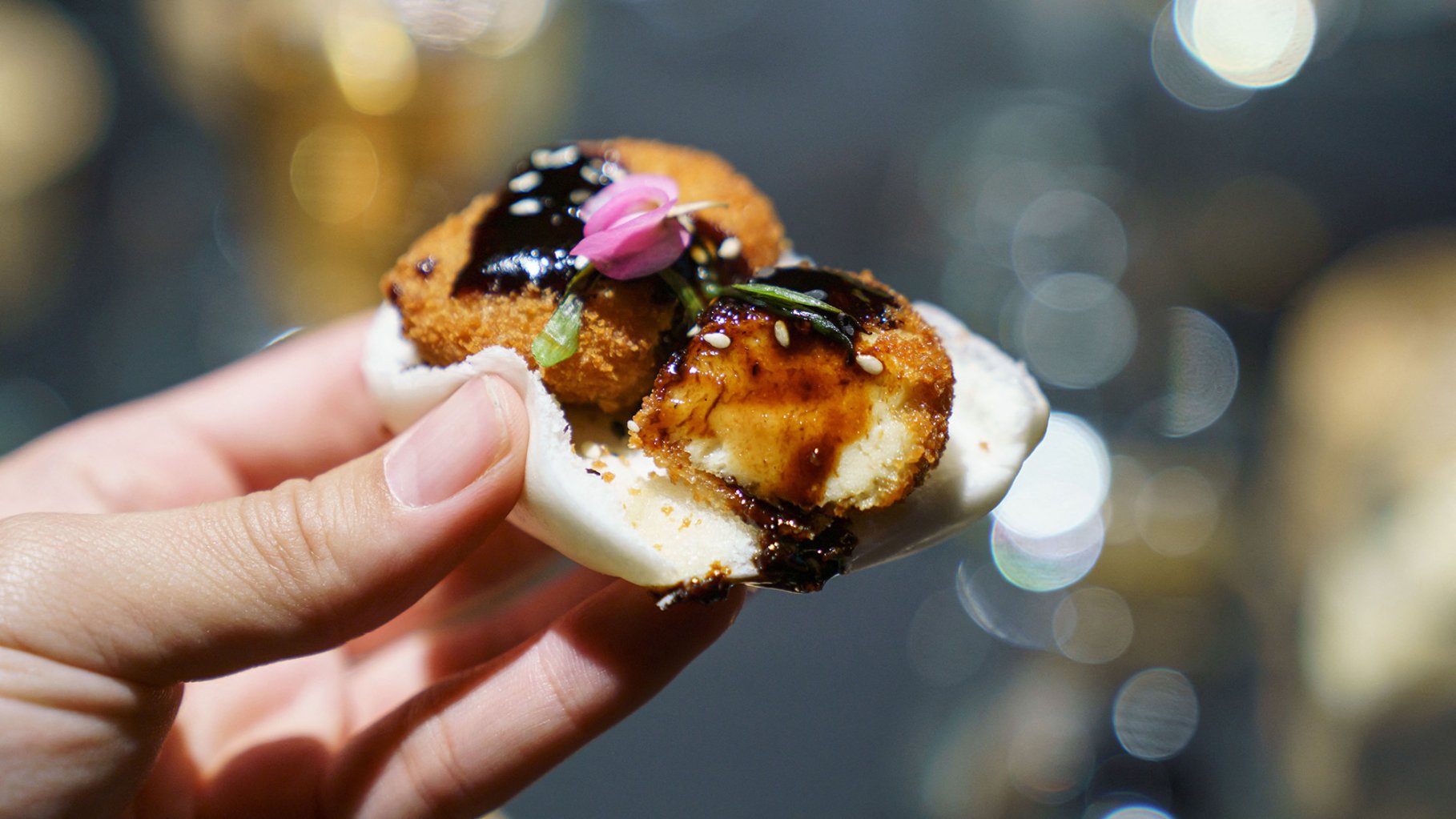 Two nuggets made from lab-grown chicken meat served on a bao with a dark sauce. This close up photograph shows a white person's hand holding the bao.