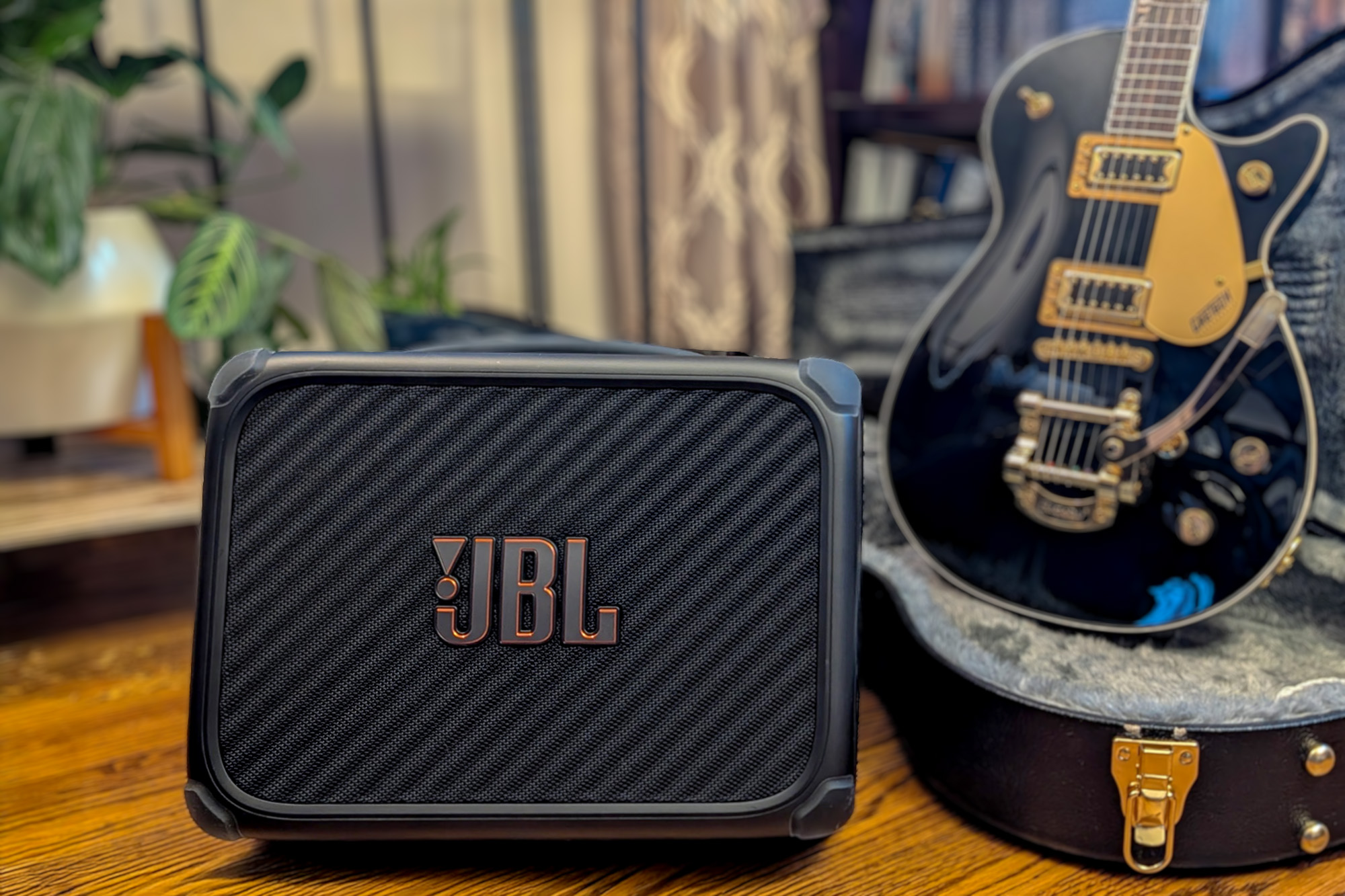A black BandBox Trio with bold JBL logo in the foreground with a Gibson guitar standing in its case behind it