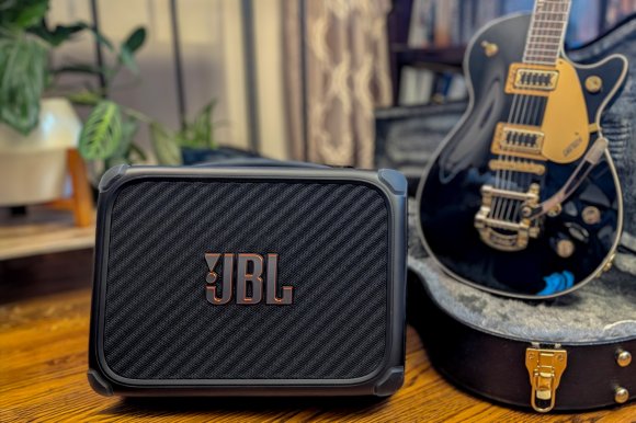 A black BandBox Trio with bold JBL logo in the foreground with a Gibson guitar standing in its case behind it