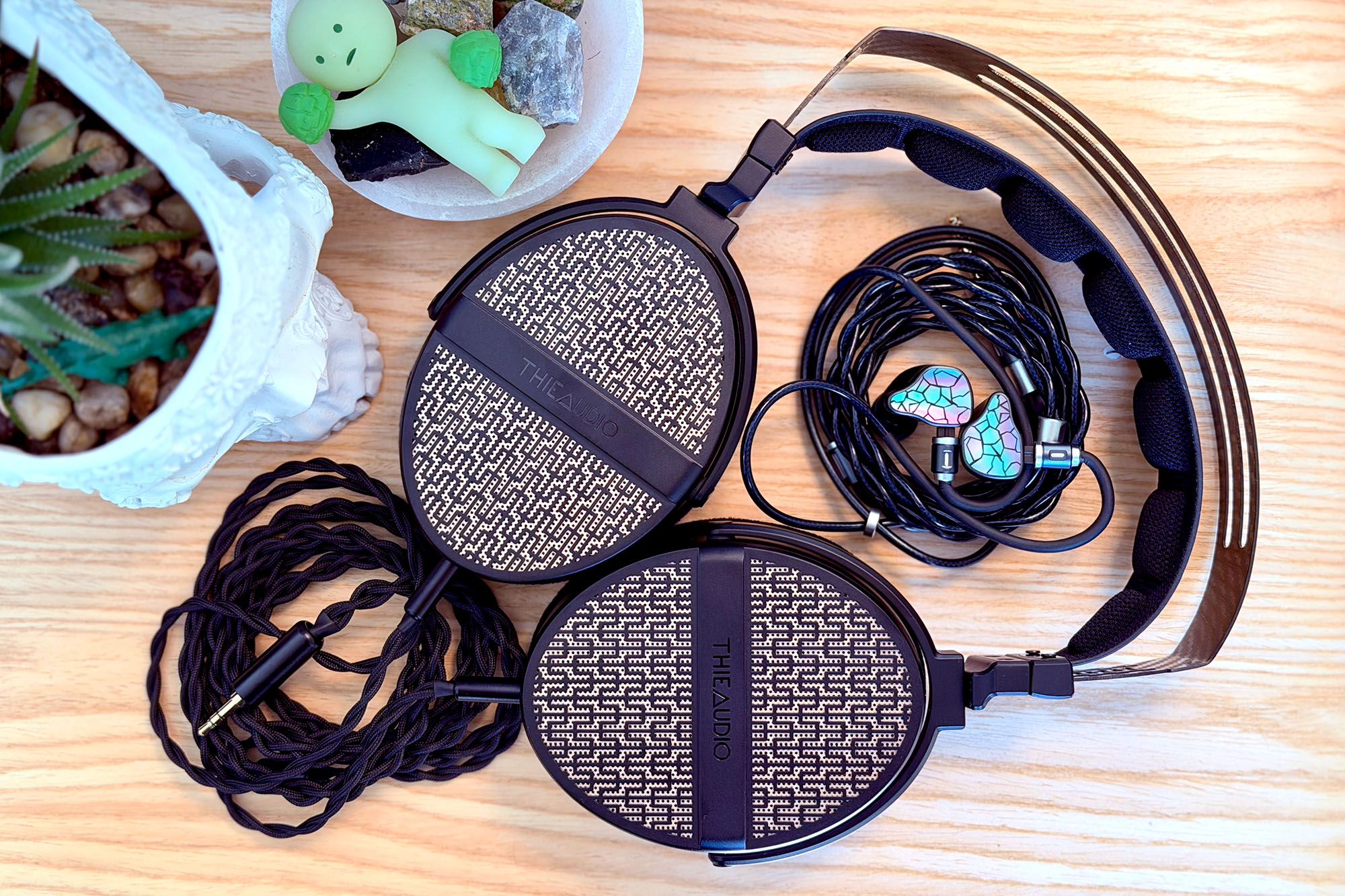 The ThieAudio Hype 4 MKII IEMs sitting inside the headband of a ThieAudio Cypher headphone laid flat on a blond wood surface with a skull planter next to it