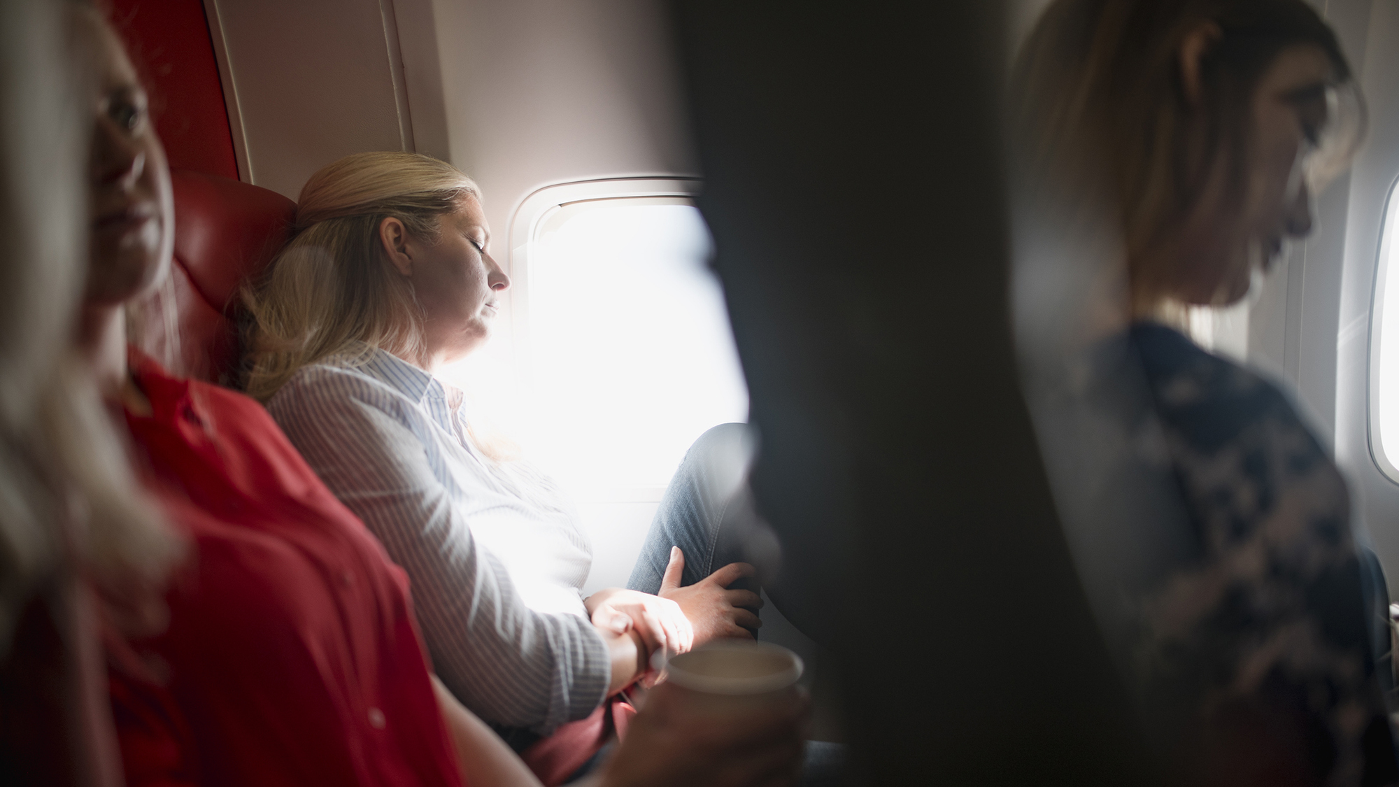 Mature woman is sleeping on a plane in economy class. She's blonde with a blue and white striped button down.