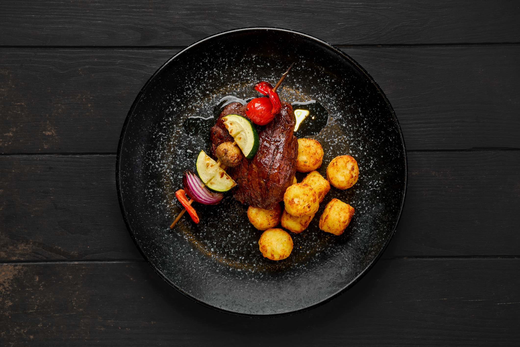 Top view of grilled beef with vegetables and potato balls on a black colored plate. The plate is sitting on a black wooden table.