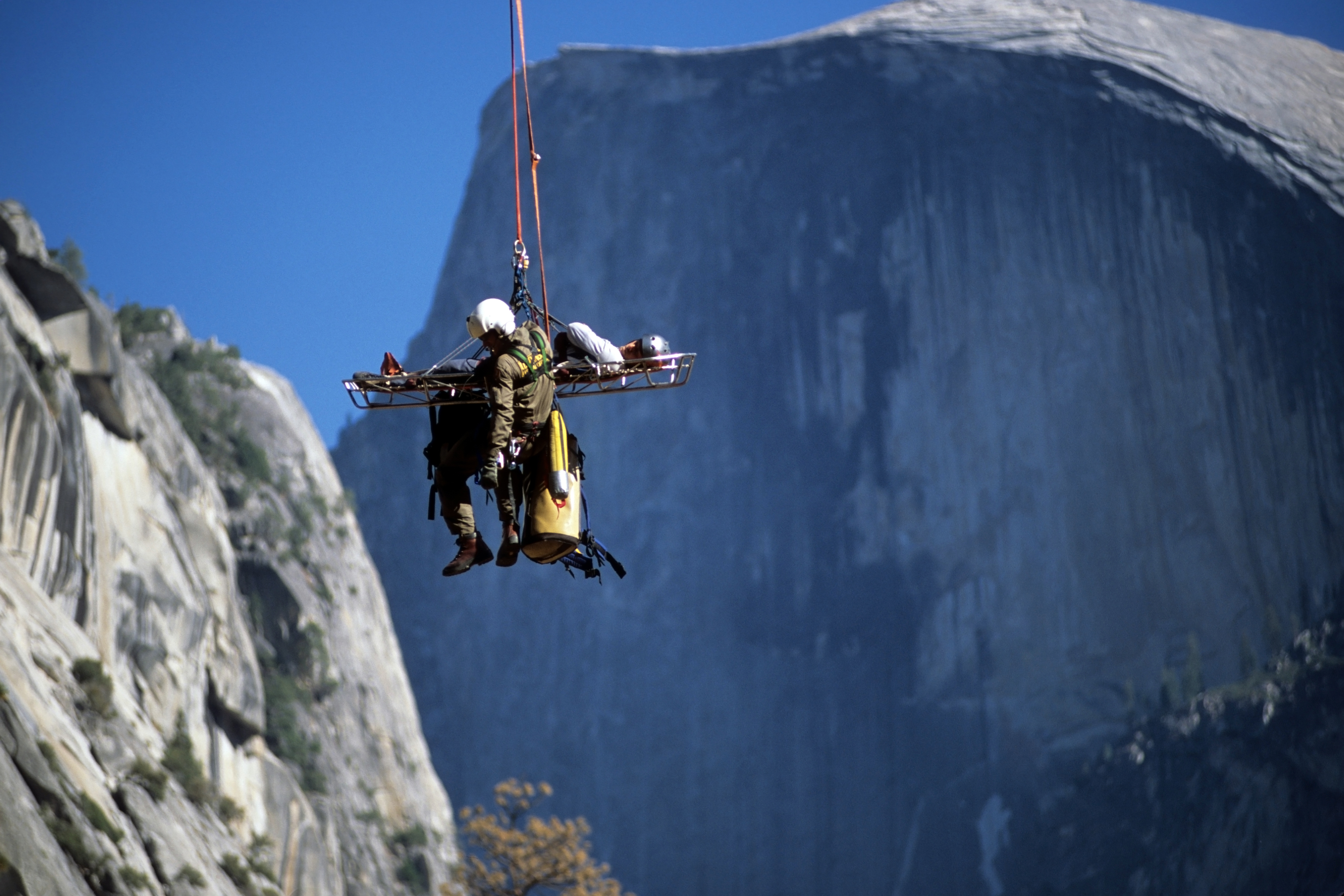 Rescuer airlifts an injured person on a gurney. We see ropes connecting them to a helicopter out of frame. Sheer cliffs are seen in the background. 
