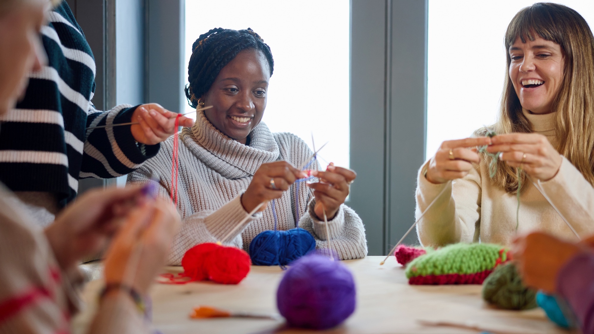 Happy group of friends enjoying craft workshop sitting at wooden table in cafe learning knitting with balls of wool, learning, togetherness, relaxation