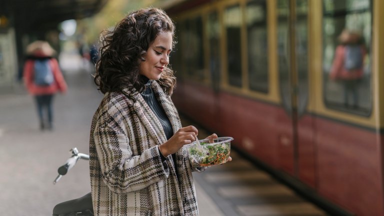 Woman having lunch on an underground station platform, Berlin, Germany. She's wearing a plaid jacket and has dark curly hair.