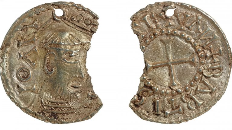 A high-resolution photograph shows both sides of a damaged, circular silver-gilt pendant depicting John the Baptist. The artifact is chipped on the right side and features a small suspension hole at the top. The obverse (left) shows a profile view of a bearded figure wearing a crown or headdress, while the reverse (right) features a central equal-armed cross enclosed in a beaded circle and surrounded by medieval-style lettering.
