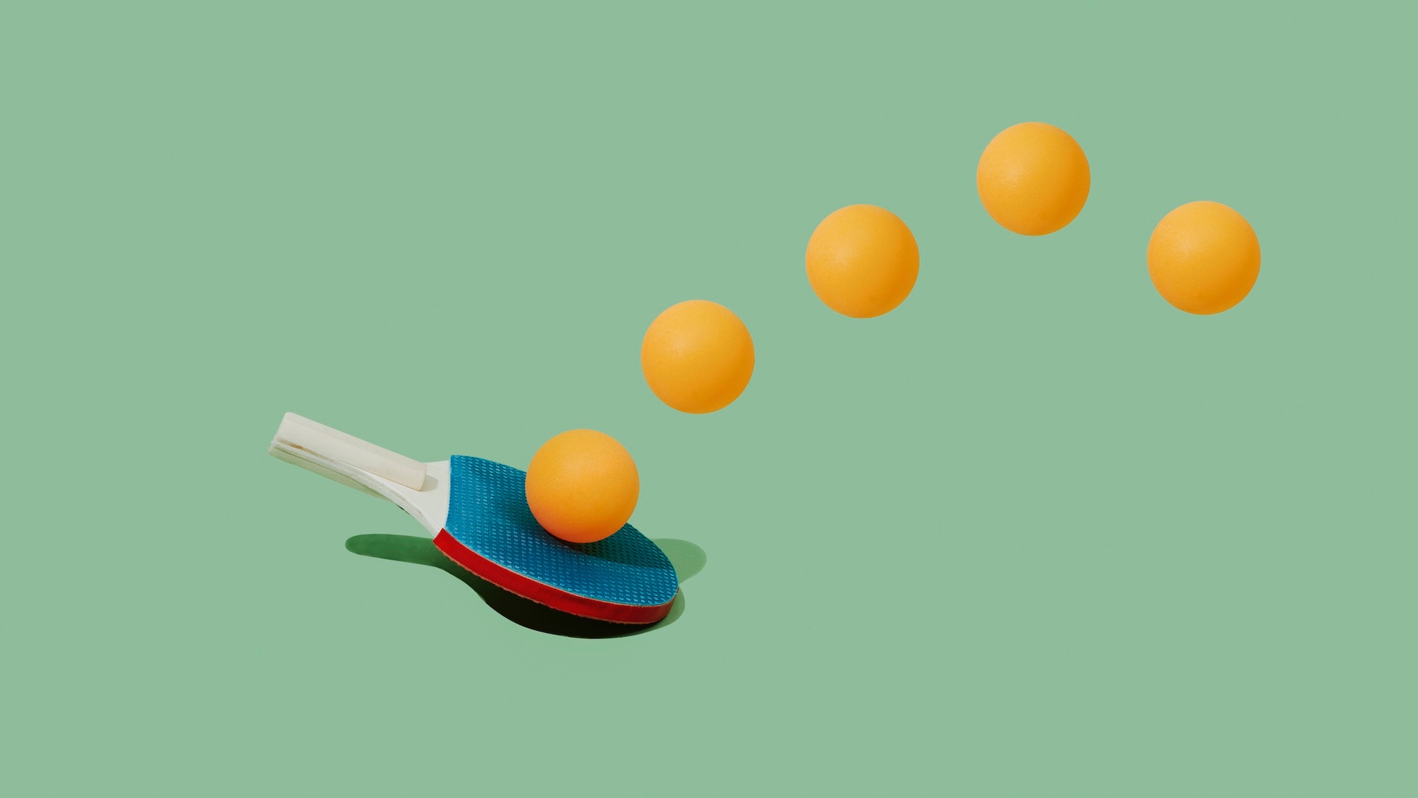 a table tennis racket and some yellow table tennis balls describing a trajectory on a pale green background
