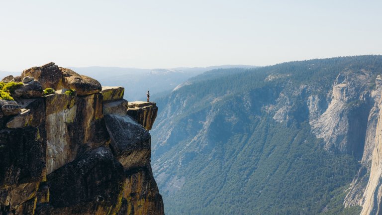 A wide-angle landscape photograph shows a lone hiker standing on the extreme edge of a sheer granite cliff, overlooking a vast mountain valley. The hiker is dwarfed by the massive, rugged rock formations and the deep, evergreen-covered canyon below. The distant mountains are shrouded in a light blue haze under a clear, bright sky, emphasizing the immense scale and potential danger of the high-altitude wilderness.