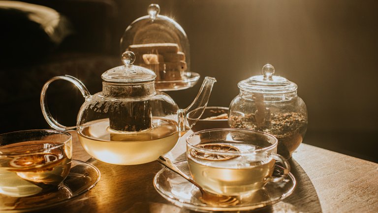 Golden light illuminates a clear teapot of green tea and two glass cups with citrus slices. A cloche of biscuits and a small bowl sit behind them, while steam and long shadows create a calm, cozy tea-time mood.
