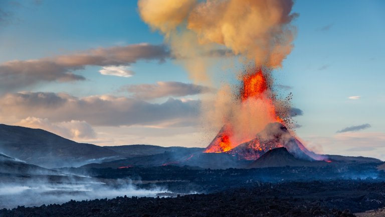 A dramatic landscape photograph depicts a fountain-style volcanic eruption at dusk. Bright orange and red lava bursts from a central vent, spraying molten rock into the air above a dark, rugged volcanic cone. A massive plume of yellow-tinted smoke and ash rises toward a sky filled with soft blue and pink clouds. In the foreground, white steam or volcanic gas drifts across a desolate, blackened lava field.