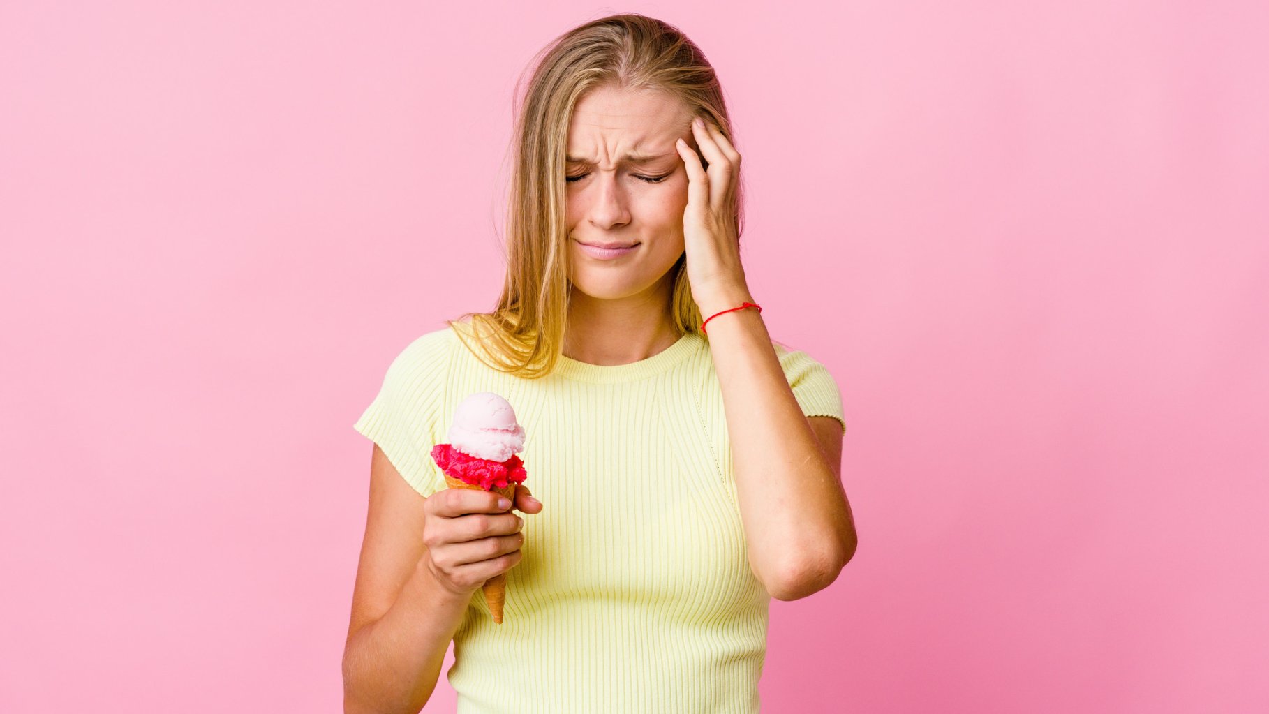 Young white woman eating an ice cream touched her temples due to a headache. She's blonde wearing a yellow shirt on a pink solid background.