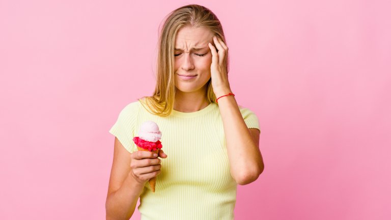 Young white woman eating an ice cream touched her temples due to a headache. She's blonde wearing a yellow shirt on a pink solid background.