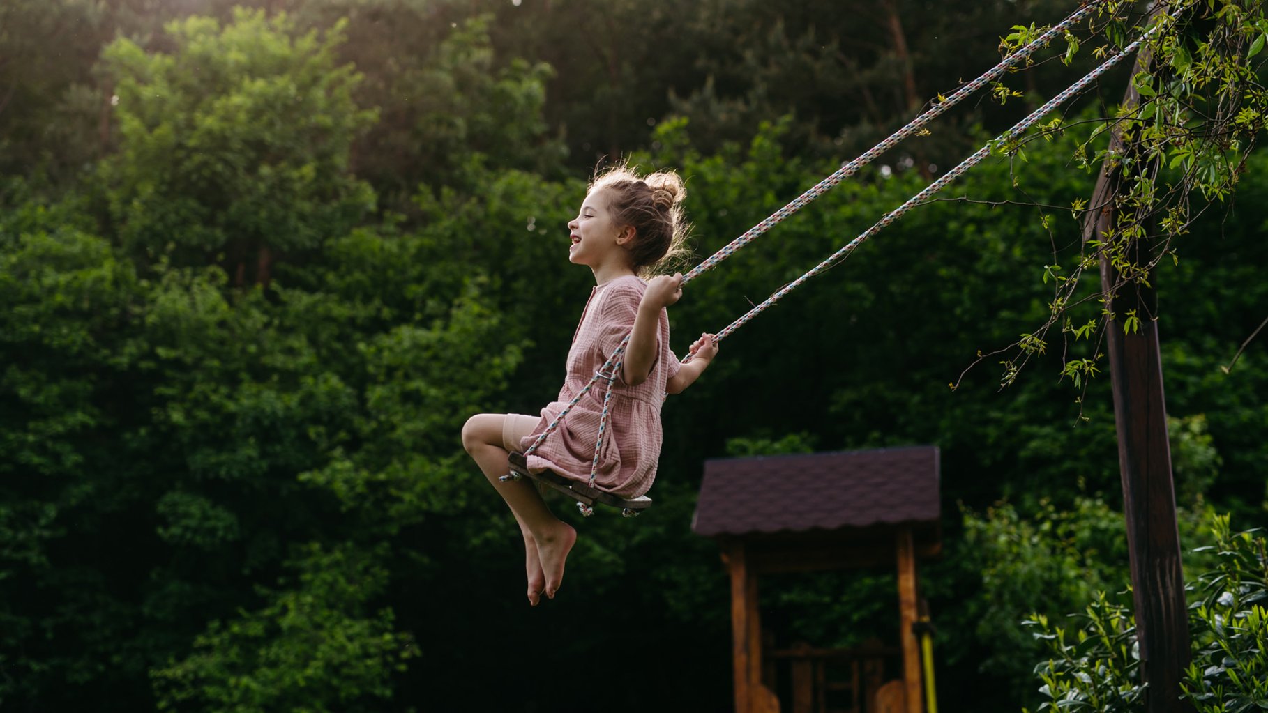 A candid, full-body photograph shows a young girl swinging joyfully in a lush, green forest setting. She is barefoot and wearing a simple pink dress, with her eyes closed and a wide smile as she glides through the air. Warm sunlight filters through the dense trees in the background, illuminating the scene and a small wooden structure behind her.
