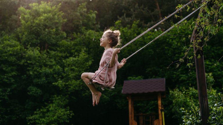 A candid, full-body photograph shows a young girl swinging joyfully in a lush, green forest setting. She is barefoot and wearing a simple pink dress, with her eyes closed and a wide smile as she glides through the air. Warm sunlight filters through the dense trees in the background, illuminating the scene and a small wooden structure behind her.