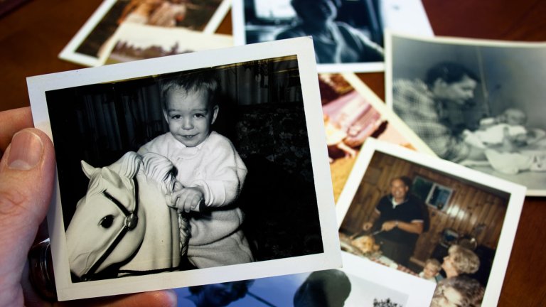 Hand holds vintage photograph of young boy riding a plastic toy horse with pile of old photos in background.