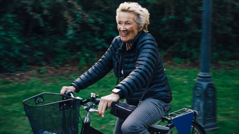 A medium-shot photograph shows a smiling senior woman with blonde hair riding a black bicycle through a park. She is wearing a black puffer jacket and grey trousers, and she is looking ahead with a joyful expression. The background is a soft blur of green grass and dark leafy bushes, emphasizing the motion of the bike.