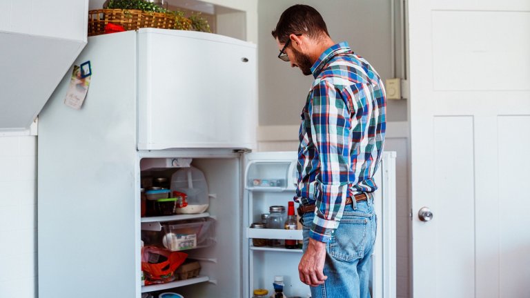 A rancher taking a break after a busy morning of work stands in his farmhouse kitchen and looks inside an open refrigerator in search of something to eat.