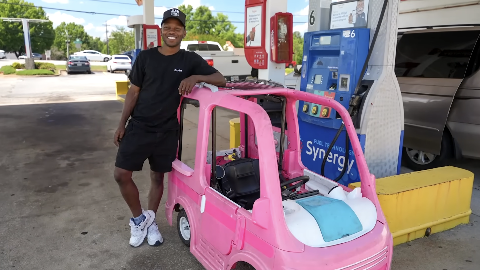 Smiling man standing next to Barbie Power Wheels Dream Camper next to gas pump at gas station